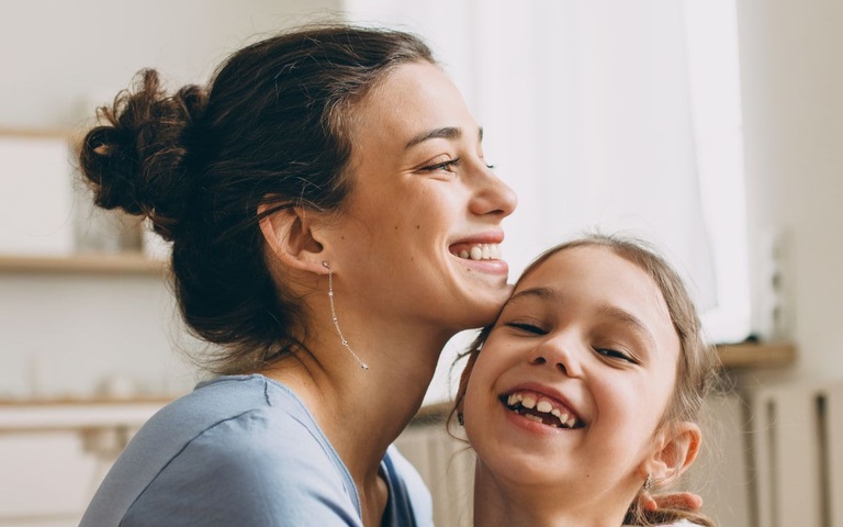 Ein kleines Mädchen sitzt lachend, mitsamt Teddybären, auf dem Schoß ihrer Mutter.